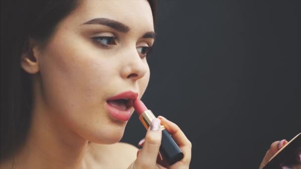 Beauty concept, head and shoulders of young woman looking at camera and painting lips with rose lipstick. Closeup portrait of model with rose lips, studio with black background. - Πλάνα, βίντεο