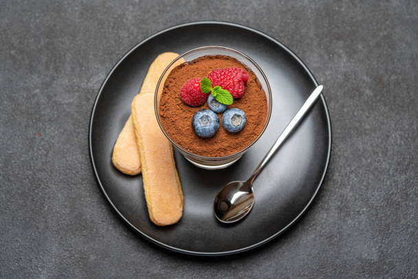 Classic tiramisu dessert with blueberries and raspberries in a glass and savoiardi cookies on dark concrete background - Foto, Imagem