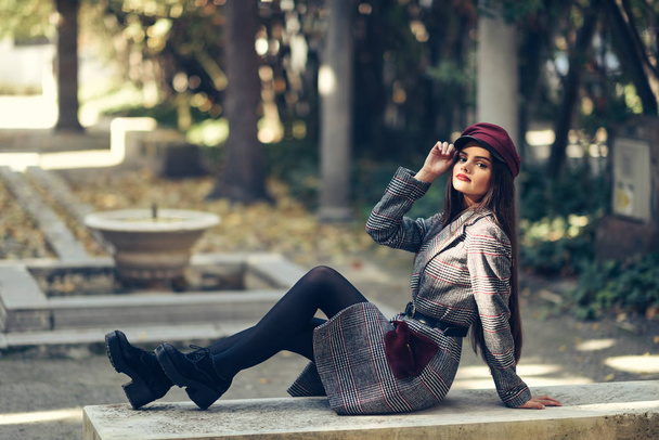 Νεαρό όμορφο κορίτσι φορώντας χειμωνιάτικο παλτό και καπάκι κάθεται σε ένα παγκάκι στο αστικό πάρκο. - Φωτογραφία, εικόνα