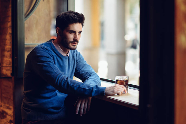 Bir soda içme bir pencere yakınında modern saç modeli ile dalgın adam. - Fotoğraf, Görsel
