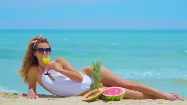 女性は海を背景に新鮮なオレンジジュースを飲みます。砂の上に海のそばに横たわっている若い美しい女性は、オレンジの新鮮なジュースを飲みます。コンセプトバケーション. - 映像、動画
