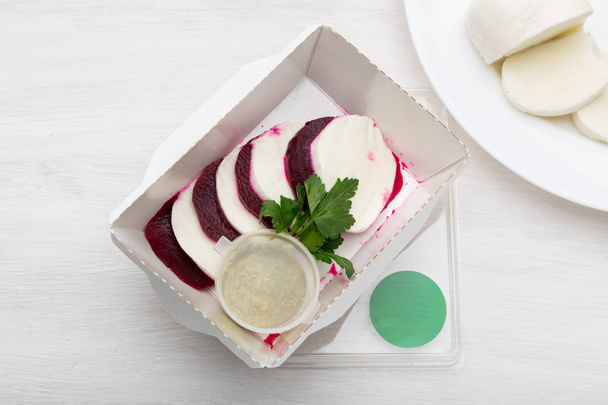 Top uitzicht van gekookte bieten met plakjes witte kaas liggen in een witte Lunchbox met zure roomsaus en peterselie op een witte tafel naast geitenkaas. Eiwit snack concept. - Foto, afbeelding