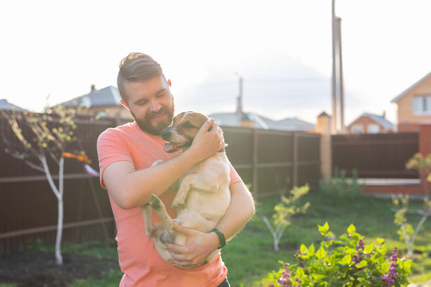 Positive young man with a beard is holding his beloved dog Jack Russell Terrier and is enjoying a joint rest in the yard of his country house on a sunny summer day. The concept of love for their four - Photo, Image