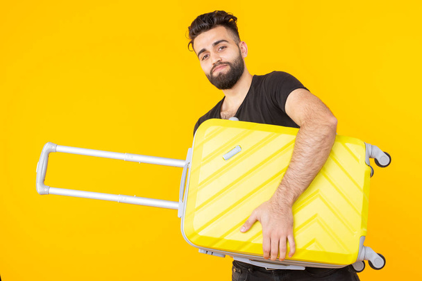 Cute pretty young arab man with a beard holding a yellow suitcase in his hands on a yellow background. Concept of travel and vacations - Photo, Image
