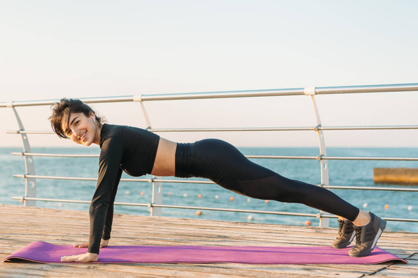 Mooie brunette jonge vrouw met witte draadloze koptelefoon in een zwarte jumpsuit neemt plank workout op een mat op de pier bij zonsopgang. - Foto, afbeelding