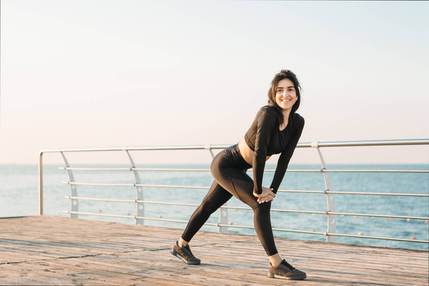 Mooie brunette jonge vrouw met witte draadloze koptelefoon in een zwarte jumpsuit neemt oefeningen op een pier bij zonsopgang kijkend naar de camera. - Foto, afbeelding