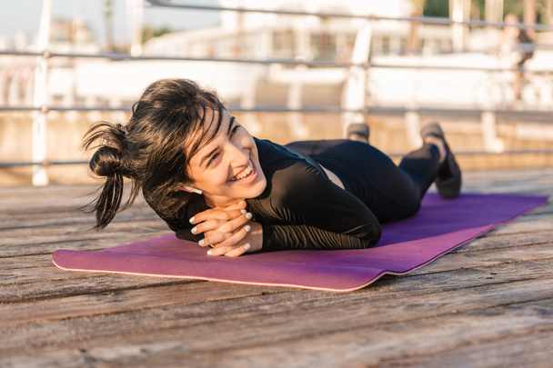 Glimlachend brunette jonge vrouw met witte draadloze koptelefoon in een zwarte jumpsuit rust na de plank workout op een mat op de pier bij zonsopgang.  - Foto, afbeelding