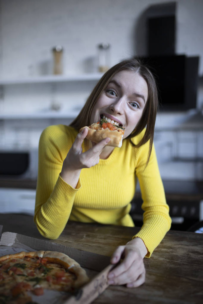 Αστείο μελαχρινή κορίτσι σε κίτρινο πουλόβερ τρώγοντας πίτσα στην κουζίνα - Φωτογραφία, εικόνα