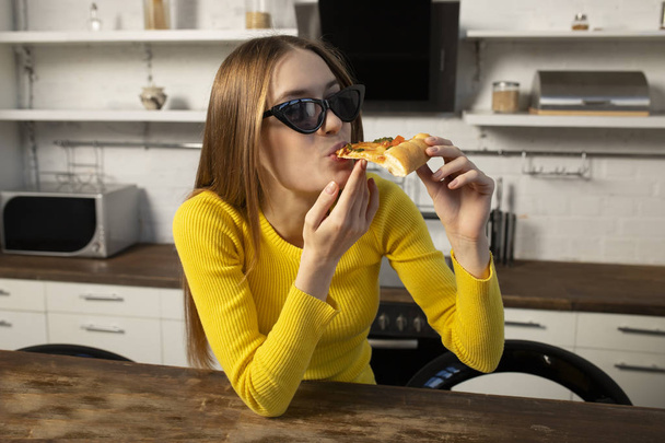 Αστείο μελαχρινή κορίτσι σε κίτρινο πουλόβερ και μαύρα γυαλιά τρώγοντας p - Φωτογραφία, εικόνα