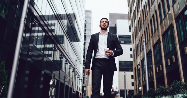 Intelligent male entrepreneur dressed in luxury suit walking near skyscrapers in financial district of megalopolis, Caucasian experienced lawyer with coffee cup going to business meeting in downtown - Φωτογραφία, εικόνα