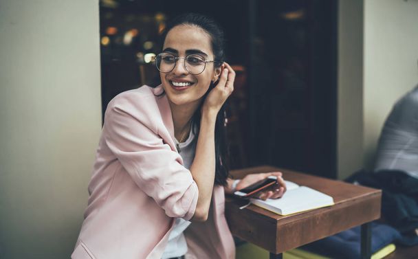 Happy female student in stylish classic spectacles laughing during time for studying in street cafeteria, positive Georgian woman in glasses using cellphone for research information for course work - 写真・画像