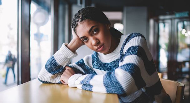 Portrait of attractive dark skinned hipster girl dressed in casual wear posing at table in university cafeteria, charming african woman looking at camera while waiting friend in cafe interior - 写真・画像