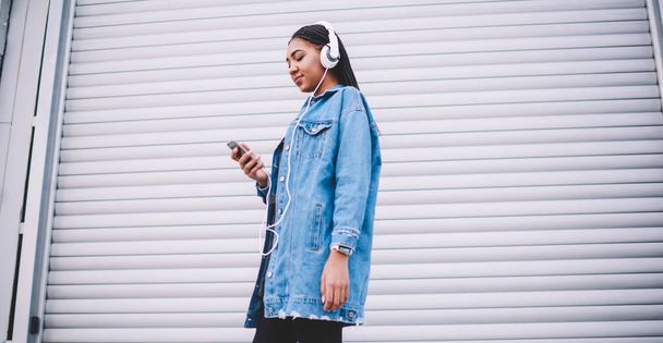 Happy millennial female meloman in earphones listening favourite playlist from multimedia application on cellphone, positive hipster girl enjoying sounds from headphones during chatting on smartphone - Foto, Imagem