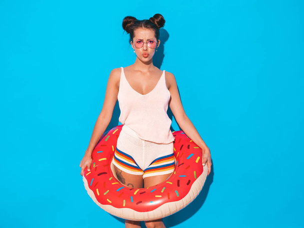 Young beautiful sexy smiling hipster woman in sunglasses.Girl in summer clothes with donut lilo inflatable mattress.Positive female going crazy.Funny model isolated on blue - Φωτογραφία, εικόνα