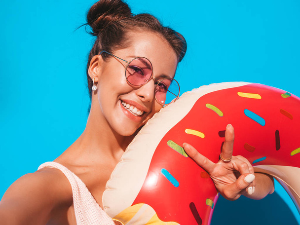 Young beautiful sexy smiling hipster woman in sunglasses.Girl in summer clothes with donut lilo inflatable mattress.going crazy.Funny model isolated on blue.Taking selfie self portrait photos on phone - Fotografie, Obrázek