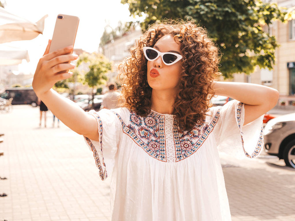 Beautiful smiling model with afro curls hairstyle dressed in summer hipster white dress.Sexy carefree girl posing in the street in sunglasses.Taking selfie self portrait photos on smartphone.Duck face - Foto, imagen