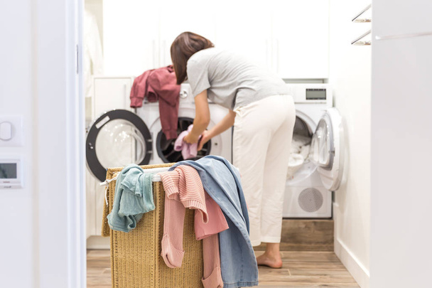 Молода жінка завантажує пральну машину і кошик повний брудного одягу в пральні
 - Фото, зображення