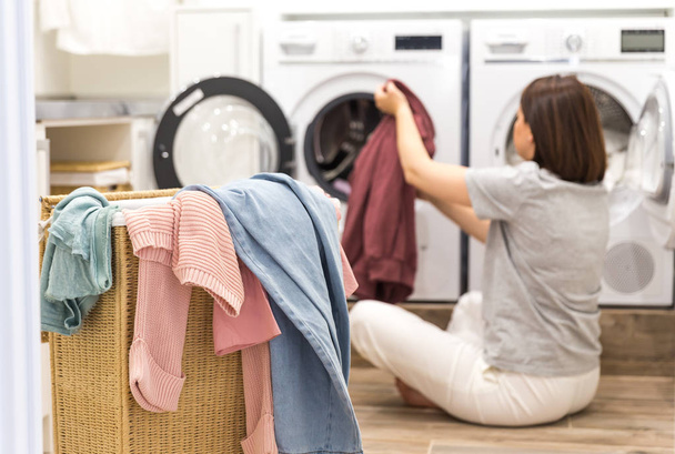 junge Frau lädt Waschmaschine und einen Korb voller schmutziger Wäsche in Waschküche - Foto, Bild