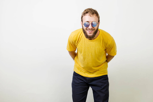 Retrato de un joven guapo con camisa amarilla, gafas de sol redondas y barba sonriente riendo mirando a la cámara sobre un fondo gris
. - Foto, imagen