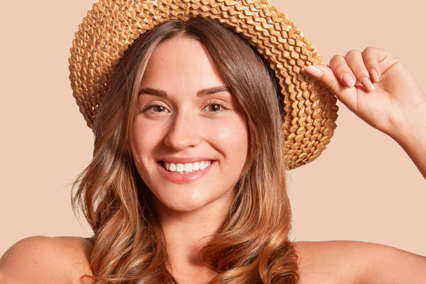 Portrait of pretty cheerful woman wearing straw hat, attractive female looking smiling directly at camera, expresses happyness, keeps hand on hat, being in good mood, posing with bare shoulders. - Photo, Image