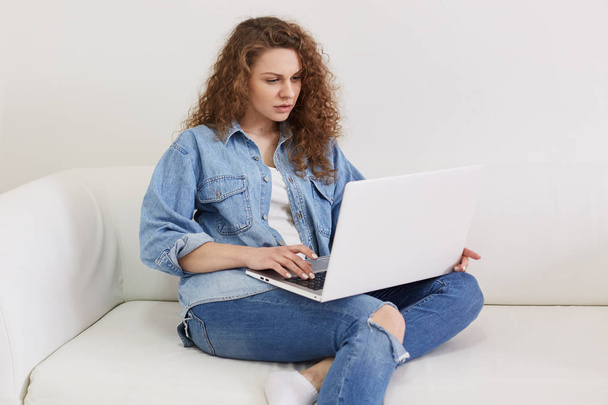 Женщина-фрилансер работает дома с ноутбуком, имеет концентрированное выражение лица, одета в джинсовую куртку и джинсы, очаровательная женщина сидит на белом удобном диване, используя беспроводной интернет
. - Фото, изображение