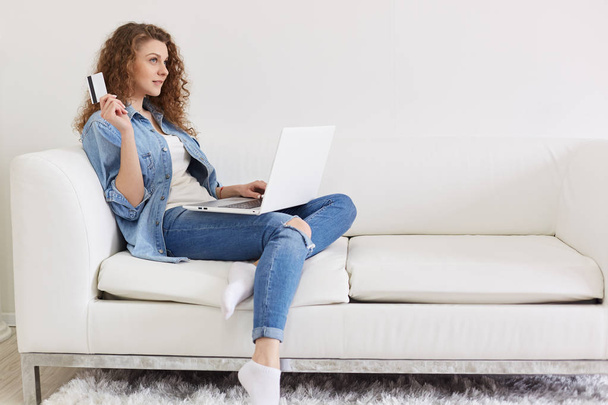 Portrait of thoughtful magnetic woman sitting with her laptop on sofa, holding credit card in one hand, thinking about purchases online, like spending money, wearing casual jeans clothes. Shopping. - Foto, Imagen