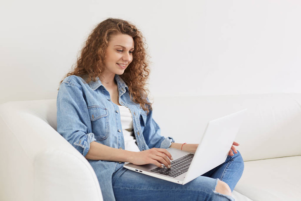 Indoor shot of positive attractive girl with curly hair chatting with her friends at social networking sites, having pleasant facial expression, looking attentively at laptop screen. Tech concept. - 写真・画像