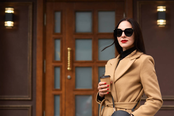 Outdoor shot of beautiful young stylish brunette woman wearing beige coat posing in open air, walks along city streets, attractive female in trendy outfit and black sunglasses, holding takeaway coffee - Фото, зображення