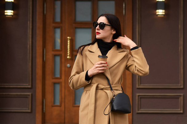 Image of stylish beautiful female standing outside, wearing beige coat, black shirt, fashionable sunglasses, touches her hair, having paper cup of coffee or tea in other hand, looking aside. - Fotoğraf, Görsel