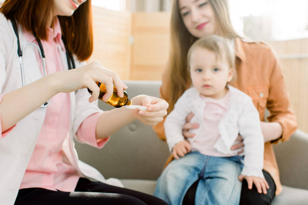 Sağlık ve tıp kavramı. Kadın el şişeden kaşık ilaç veya antipiretik şurubu dökme. Küçük bebek kız ve güzel anne arka planda kanepede oturan - Fotoğraf, Görsel