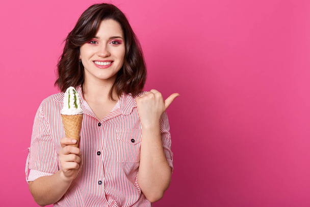 Indoor studio shot of positive beautiful young woman standing isolated over pink background, holding ice cream in one hand, making gesture with thumb, smiling sincerely. Copy space for advertisement. - 写真・画像