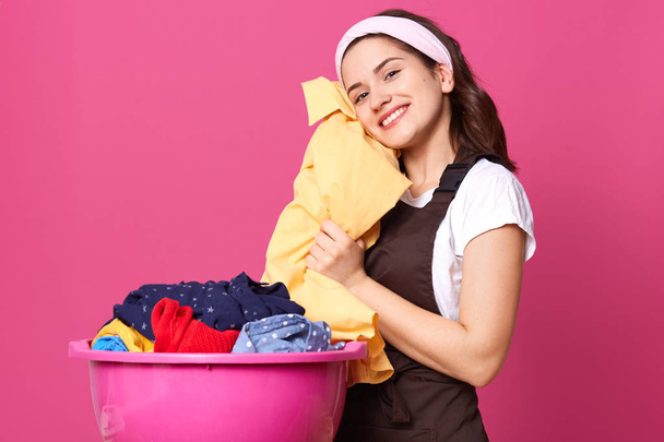 Indoor studio shot of adorable European housewife putting fresh clothes close to face, having toothy smile on face, coping with household chores, getting pleasure, wearing apron, t shirt and headband. - Foto, imagen