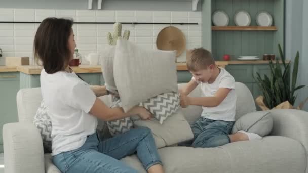 Son and mom playing together on bed at home. Little cute boy having funny pillow fight with mom on couch in living room. Loving mother spending time with her son. Family in white shirts and blue jeans - Imágenes, Vídeo