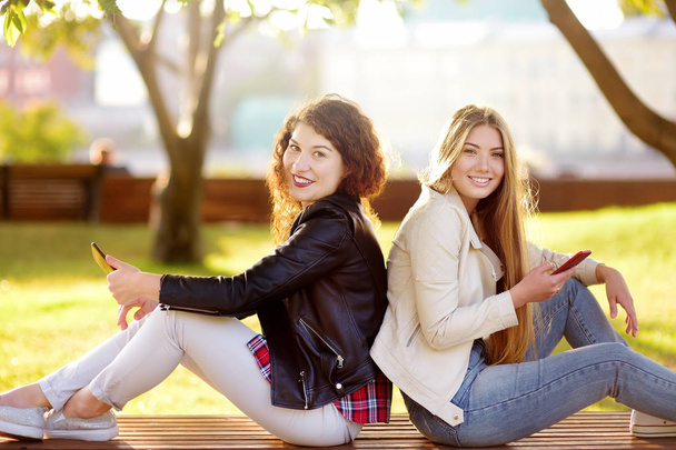 Two beautiful young women talking while sitting on a bench at sunny park. - Foto, Bild