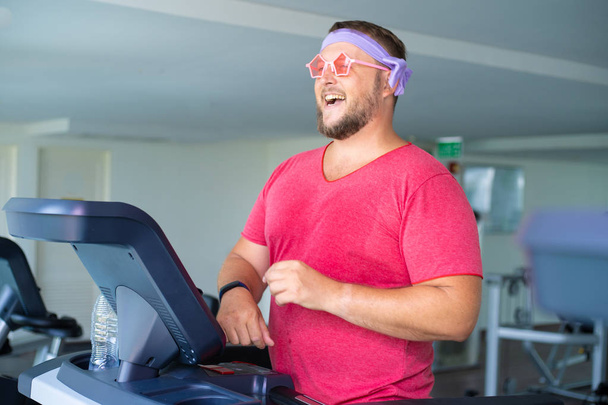 Playful fat man in a pink T-shirt and pink glasses is engaged in fitness in the gym. - Photo, Image