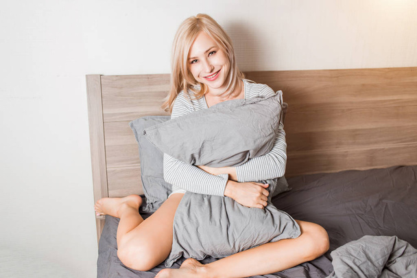 Позитивна жінка з подушкою сидить у ліжку і посміхається до камери вдома
 - Фото, зображення