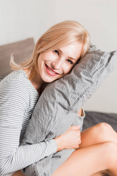 Positive woman with pillow sitting in bed and smiling to camera at home - Фото, зображення