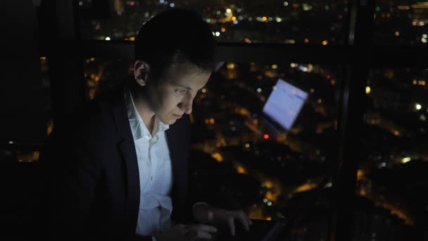 Ο νεαρός ελεύθερος επαγγελματίας δουλεύει στον υπολογιστή του κοντά στο παράθυρο στο σπίτι τη νύχτα.. - Πλάνα, βίντεο