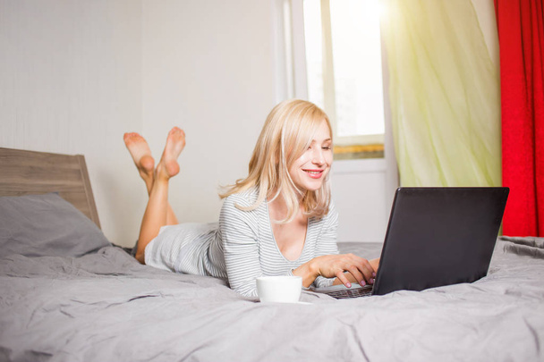 Pretty woman using laptop while having a cup of coffee on bed - Foto, Bild