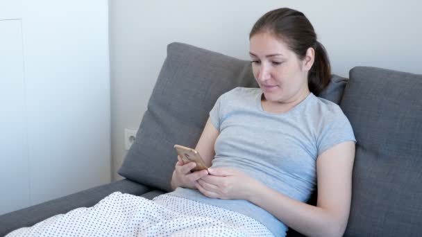 Woman is watching something funny on mobile phone sitting on the sofa. - Video, Çekim