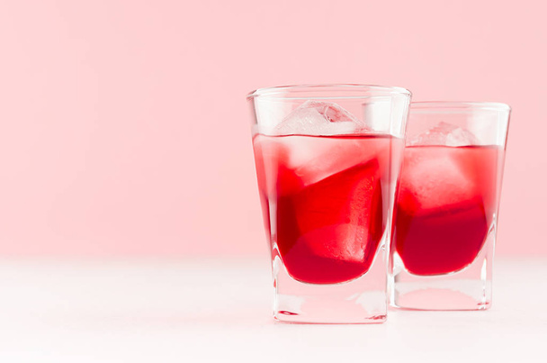 Summer fresh red strawberry drinks with ice cubes in two shot glasses on fashion pastel pink color background and white wood table, copy space. - Foto, afbeelding