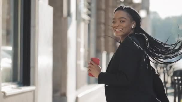 Young happy African American businesswoman in earphones walking the city street while listening to the music on the smartphone and dancing. Outdoor. - Footage, Video