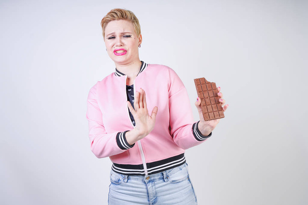 plus size caucasian girl with short haircut stands in pink sports jacket with chocolate bar in her hands and shows hate emotions for sweets. young woman wants to be healthy and slim. diet concept. - Photo, Image