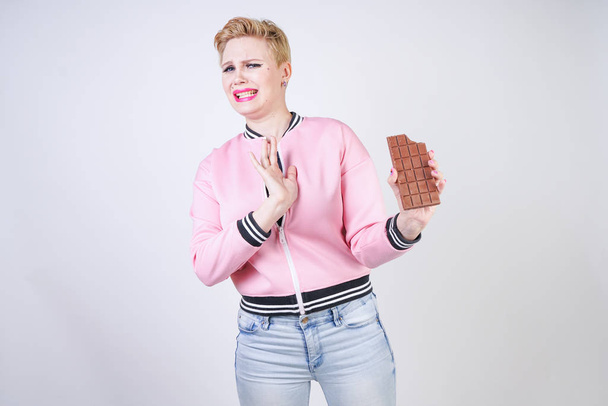 plus size caucasian girl with short haircut stands in pink sports jacket with chocolate bar in her hands and shows hate emotions for sweets. young woman wants to be healthy and slim. diet concept. - Фото, изображение