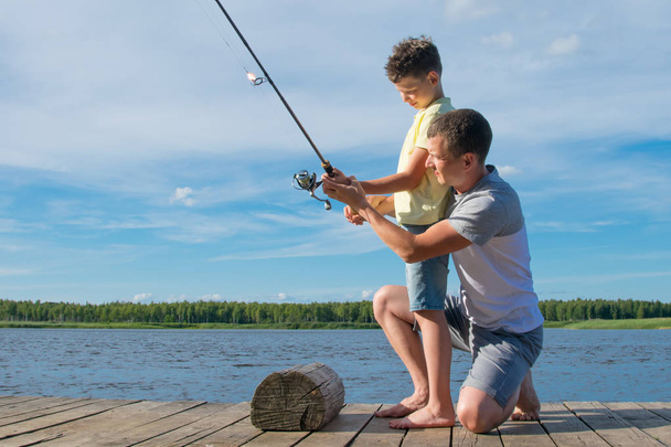 father shows his son on the pier how to hold a fishing rod to catch fish, against a beautiful landscape - Foto, Bild