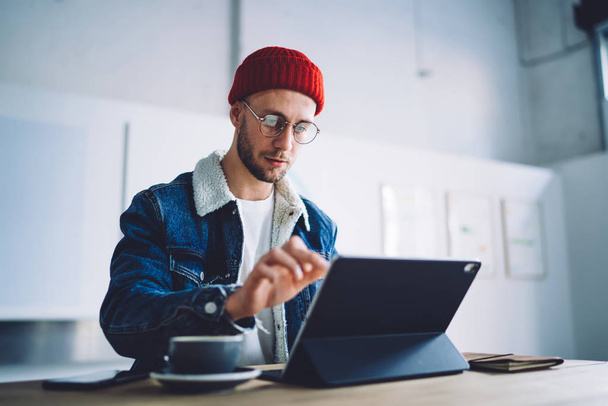 Millennial man in optical spectacles for provide eyes protection typing on graphic tablet touchscreen for updating communication and connection settings, clever male freelancer searching information - Foto, Imagem
