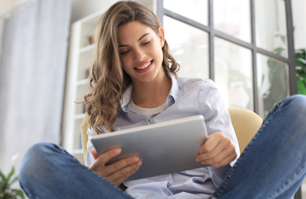 Smiling young female sitting in the armchair in the living room and using with her tablet. - Foto, Imagen