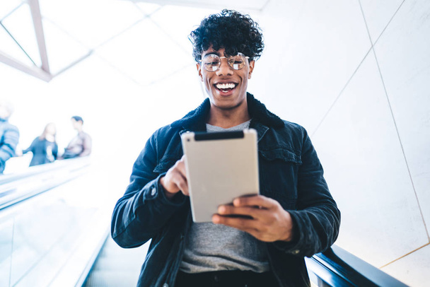 Cheerful Latino man in classic trendy glasses for vision correction going down at escalator and feeling excited from online chatting via modern touch pad, millennial happy male blogger reading sms - Fotografie, Obrázek
