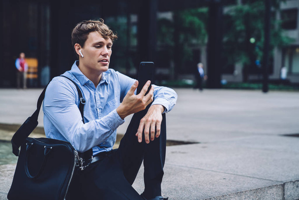 Caucasian businessman in formal wear making international cell conversation with partner while sitting at city area of financial district, experienced male trader in headphones watching video - Photo, Image