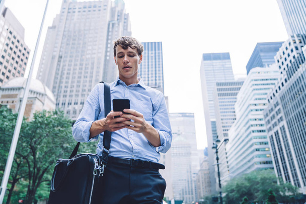 Below view of young employer in formal wear reading sms message on modern mobile phone standing at urban setting in downtown and using 4g wireless internet connection for communicate with colleagues - Fotó, kép